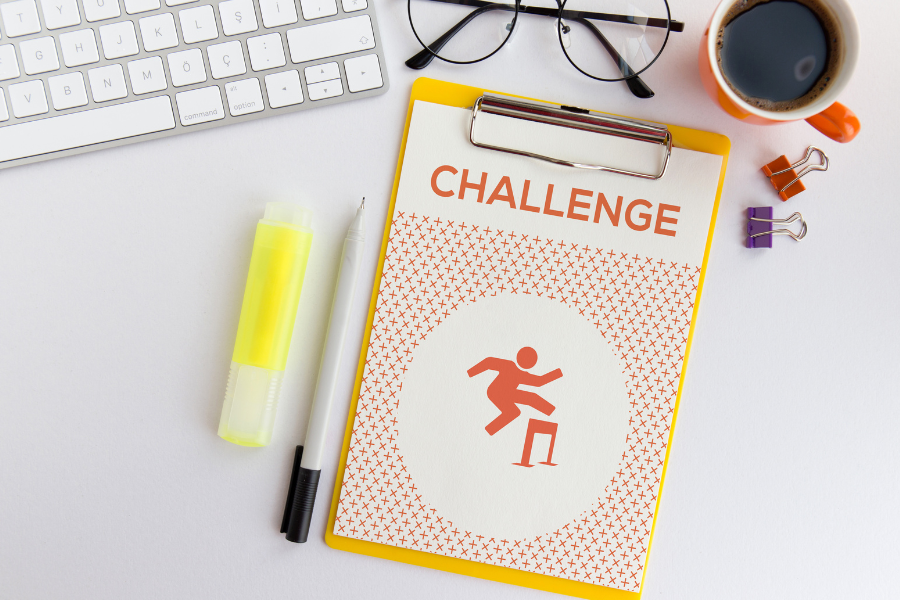 A desk with a keyboard, spectracles, mug of coffee, bulldog clips, a yellow highlighter, a pen and a clipboard with a piece of paper with the heading 'Challenge' written on it and below that an orange stick figure jumping over a hurdle