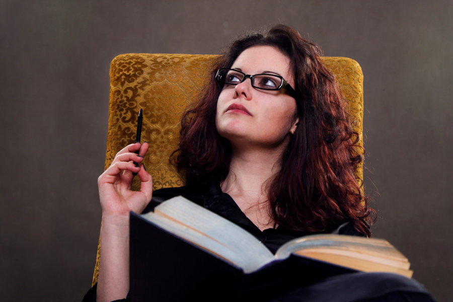 A white woman with long dark wavy hair and glasses sitting in a mustard-colour chair holding a pen and a big notebook.