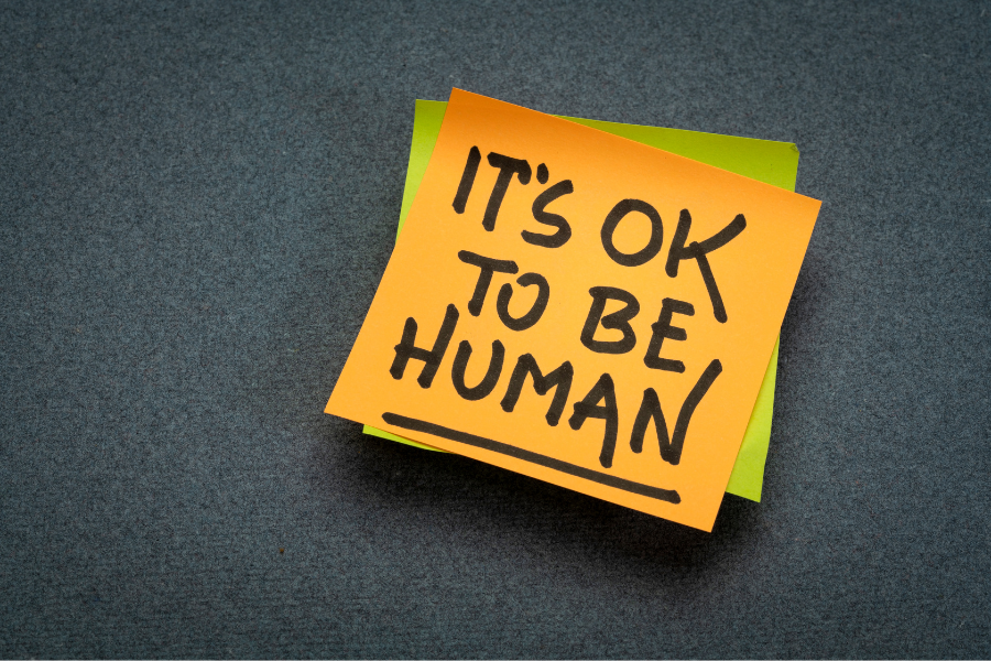 A green post it note with a yellow post it note on top of it with 'IT'S OK TO BE HUMAN' written on it in black capital letters